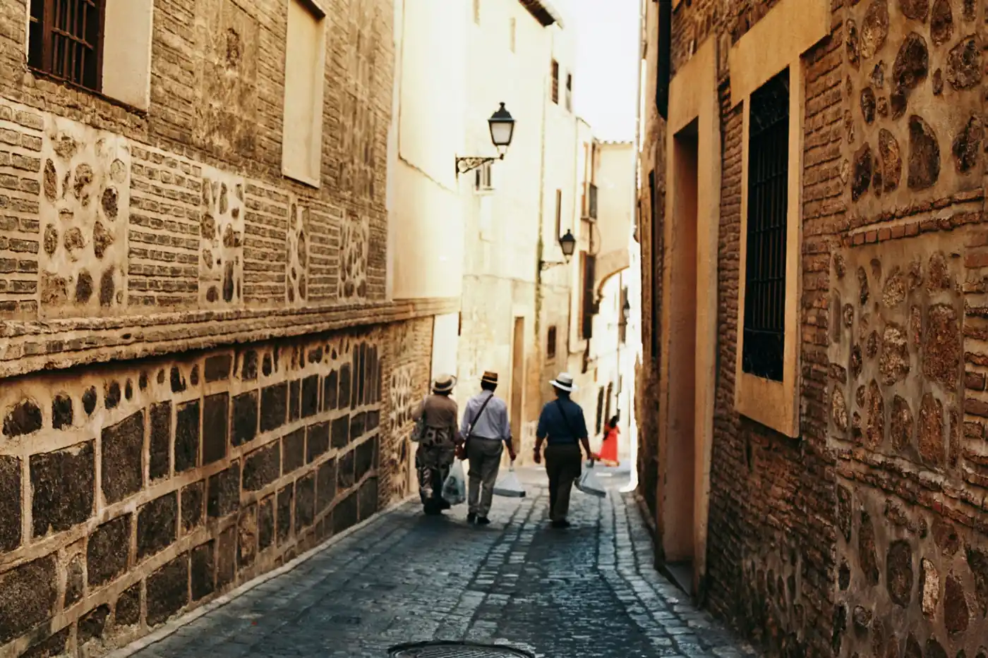 Visitors walking down a narrow cobblestone street lined with ancient stone walls in Toledo's historic Jewish Quarter (Judería)